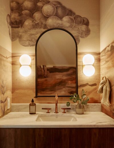 dimly lit bathroom with artistic illustration of desert as wallpaper, geometric mirror, round wall sconces, undermounted sink, has a western surreal feel