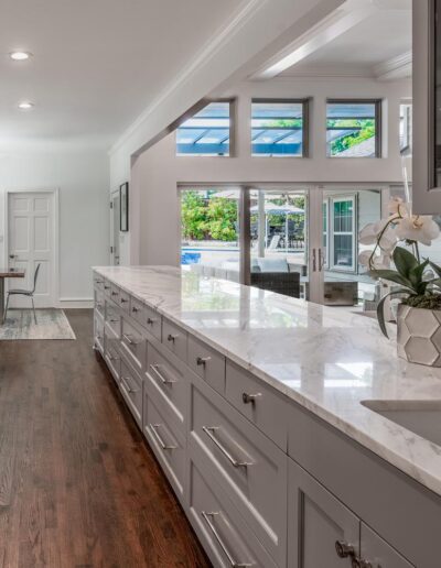 an all white kitchen with an expansive island, barstool chairs, gorgeous marble countertops, under mount sink, modern shaker cabinets with silver pulls, and a wooden floor. Next to the kitchen are large windows looking out towards the backyard