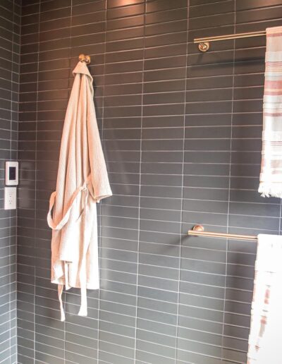 Primary bathroom wall with built-in wood cabinetry, dark subway tile, two towel racks, and a wall hook holding a robe