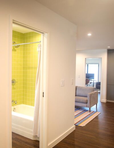 Interior hallway with flat white walls, wood floors, and simple baseboards leading to a contemporary bathroom with bright green shower tiles and a sitting area with contemporary floor-to-ceiling storage cabinets