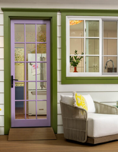 A cozy patio with white cushioned chairs, a small table with mugs, a blue planter with yellow flowers, and a glass-paneled lavender door next to a green-trimmed window set in cream siding.