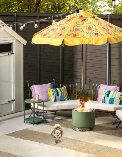 A cozy backyard patio features a white shed, curved benches with colorful pillows, a fire pit, a yellow floral umbrella, string lights, a potted plant, and a small dog standing on a rug.
