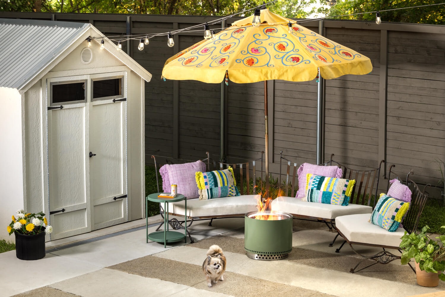 A cozy backyard patio with a white shed, string lights, colorful cushions on white outdoor furniture, a yellow umbrella, a green fire pit, potted flowers, and a small dog standing on a rug.