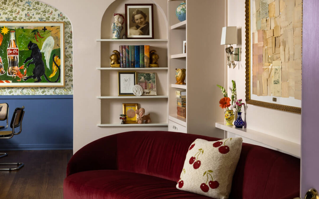 A cozy, stylish living room with a curved burgundy sofa, cherry-patterned pillow, patterned rug, built-in shelves with books and art, and framed artwork on pastel walls. A doorway leads to an adjacent room.