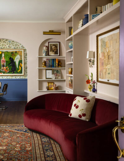 A cozy, stylish living room with a curved burgundy sofa, cherry-patterned pillow, patterned rug, built-in shelves with books and art, and framed artwork on pastel walls. A doorway leads to an adjacent room.