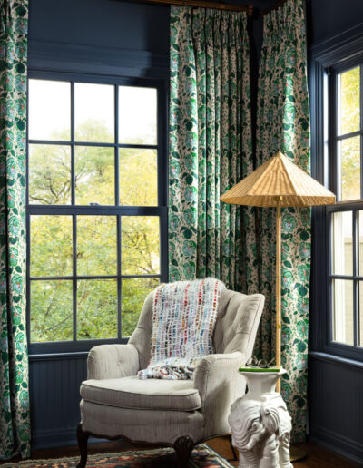 Cozy reading nook with a light armchair draped in a colorful blanket, next to an elephant-shaped side table and a standing lamp with a woven shade. Green floral curtains frame large windows showing trees outside.