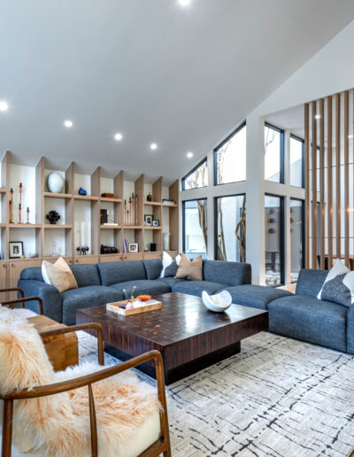 Modern living room with gray sectional sofa, wooden coffee table, armchairs with fur throws, open shelving along the wall, decorative items, and large windows letting in natural light. Dining area is visible in the background.