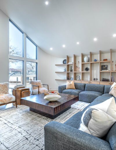 Modern living room with large windows, a grey sectional sofa, a wooden coffee table, two armchairs, an area rug, open shelving with decor items, and a wooden sideboard with a lamp.