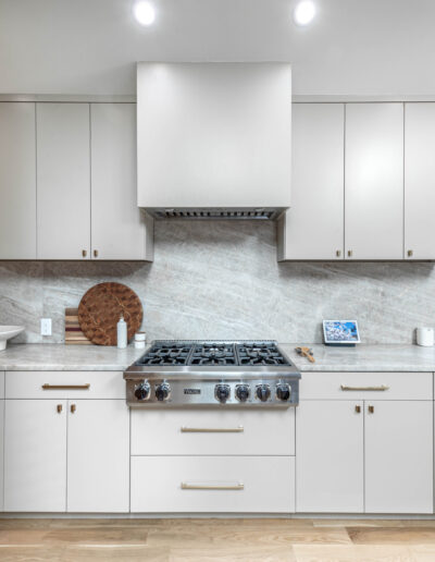 Modern kitchen with light gray cabinets, stainless steel gas stove and range hood, marble backsplash and countertops, built-in seating area on the right, and light wood flooring. Kitchenware and decor sit on the counters.