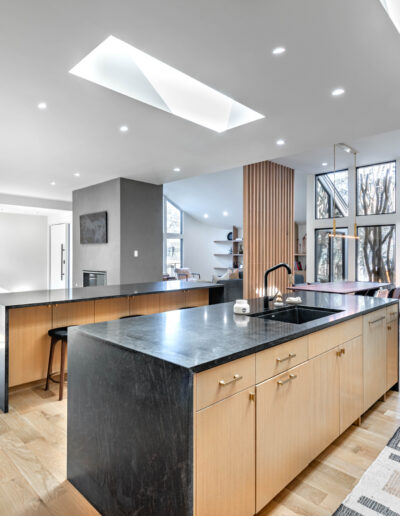 Modern open-concept kitchen and living area with light wood floors, black countertops, large island with sink, skylights, and floor-to-ceiling windows letting in natural light. A colorful painting hangs in the living space.