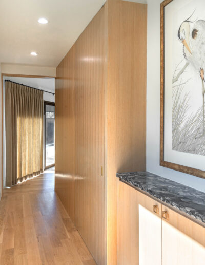 A hallway with light wood floors and paneling, a black marble-topped cabinet, a large framed bird illustration on the wall, and sunlight streaming in through a window with gold curtains at the end.