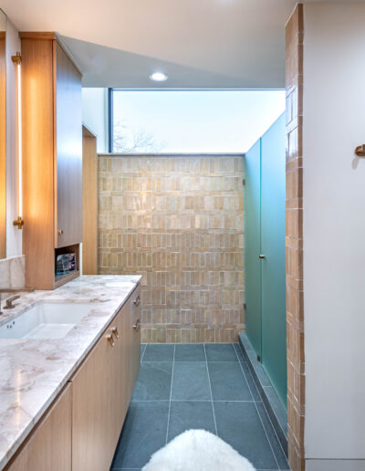 Modern bathroom with double sinks, marble countertop, light wood cabinets, large mirror, walk-in shower with tan tiles and a glass panel, gray tile floor, and a colorful robe hanging on a wall hook.