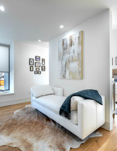 Bright modern bedroom with a white chaise lounge on a cowhide rug, abstract art on the wall, and a cozy bed visible in the background. Large window with a shade lets in natural light.