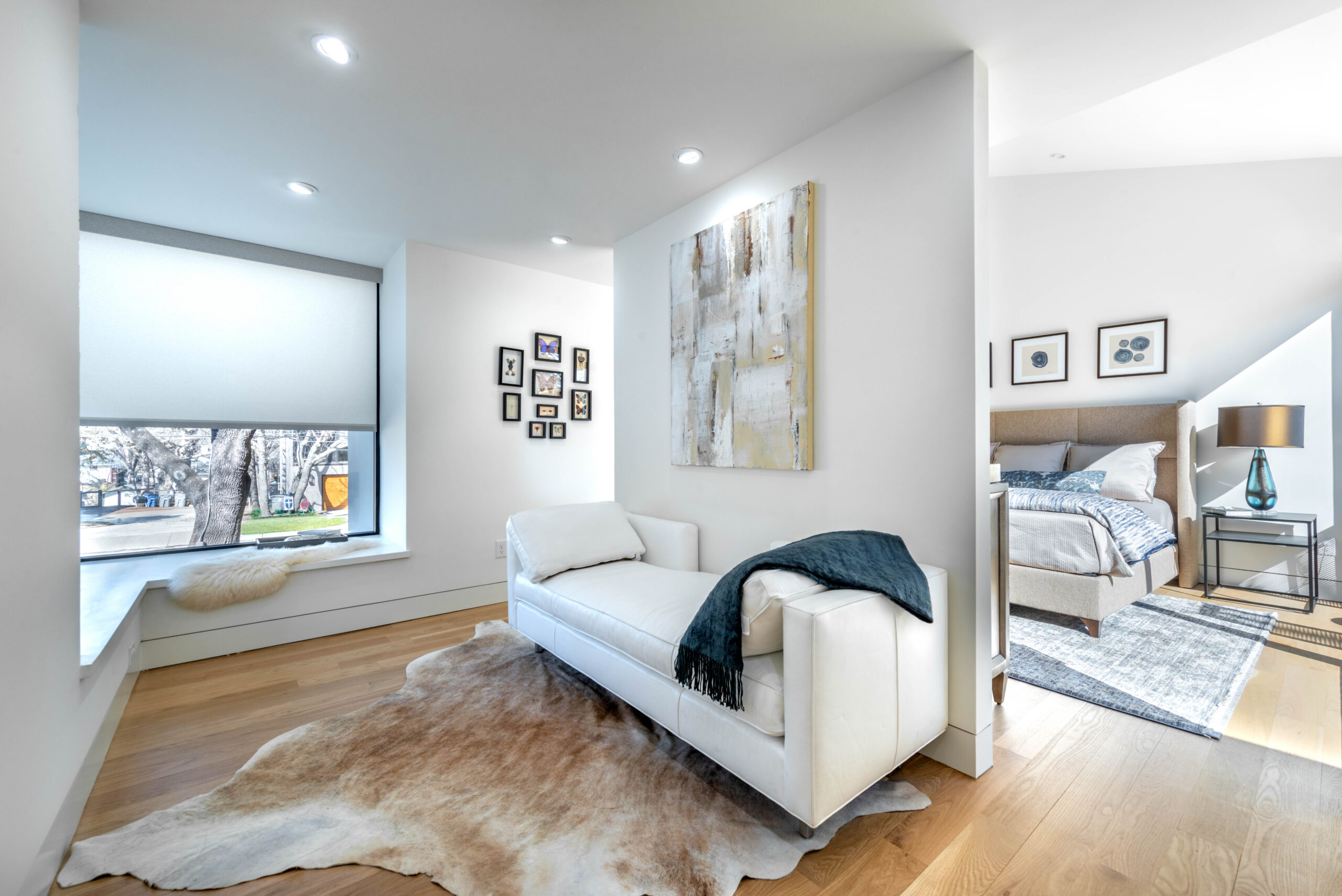 Bright modern bedroom suite with a chaise lounge on a cowhide rug, abstract art on the wall, framed photos, large window with a tree view, and a cozy bed with neutral bedding and a blue throw.