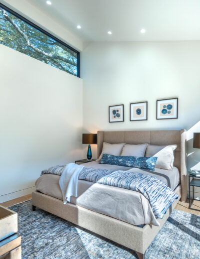 Bright, modern bedroom with large windows, neutral-colored bed, two blue glass lamps on side tables, framed art above headboard, a blue patterned rug, and an open view into a bathroom.
