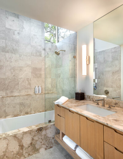 Modern bathroom with a marble-tiled shower and bathtub combination, a large vanity with a marble countertop, wood cabinetry, two vertical wall lights, and a wide mirror above the sink. Natural light enters from a window above.
