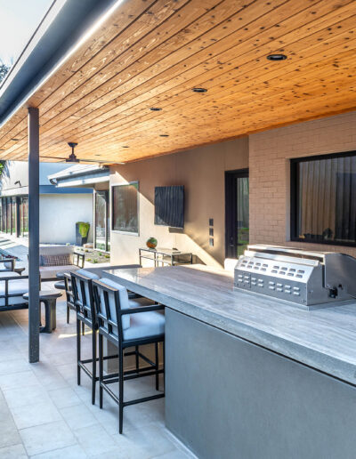 Modern outdoor kitchen and patio area with a built-in grill, bar seating, stone countertops, wooden ceiling, and lounge seating along a beige brick wall, surrounded by landscaping and greenery.