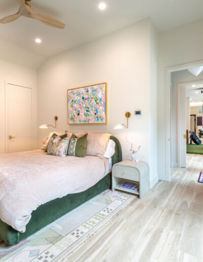 A modern bedroom with a large bed, pastel bedding, green bed frame, abstract wall art, two bedside tables, wall-mounted lamps, light wood floors, and an open door leading to a bright hallway with exercise equipment visible.