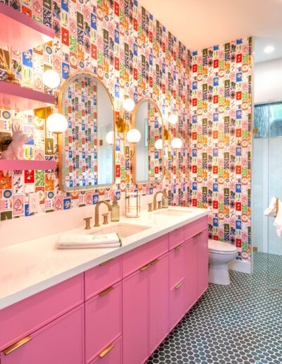Colorful bathroom with patterned wallpaper featuring various designs, a double sink vanity with pink cabinets, round mirrors, globe lights, open pink shelves with towels, and a glass shower at the back.