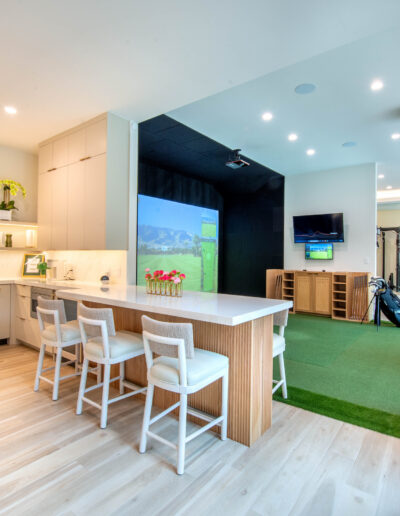 A modern home gym with a golf simulator, putting green, TV, aerial yoga swing, and a kitchenette with barstools and open shelving. The space has light wood floors and bright lighting.