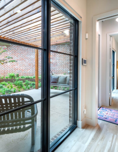 Bright hallway with light wood flooring leads to a cozy room with a pink chair and lamp. Large glass door opens to a covered patio with chairs and greenery, framed by a brick wall and wooden pergola overhead.