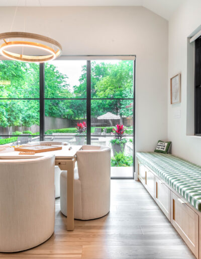 A modern dining area with a light wood table, six rounded white chairs, a green-checkered cushioned bench, large windows, and a view of a lush garden outside.
