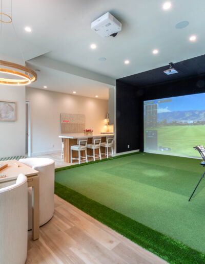 A modern recreation room with a golf simulator, green turf flooring, a golf bag, a seating area with round chairs and a table, a bar with stools, and a large screen displaying a golf course.