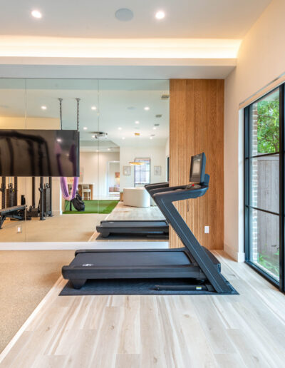A modern home gym with large windows, treadmill, aerial yoga swings with purple fabric, free weights, mirrored wall, TV, and view of greenery outside. Light wood floors and bright lighting create a spacious feel.