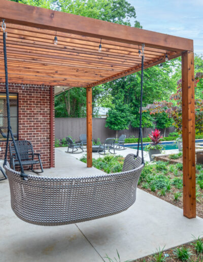 A backyard patio with a wooden pergola, a woven hanging bench swing, two rocking chairs, string lights, landscaped garden beds, and a view of more chairs and greenery beyond the patio area.