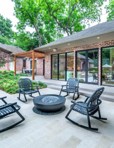 Modern brick patio with black rocking chairs surrounding a round fire pit, large glass doors leading inside, lush greenery, and a pergola covering an outdoor seating area in the background.