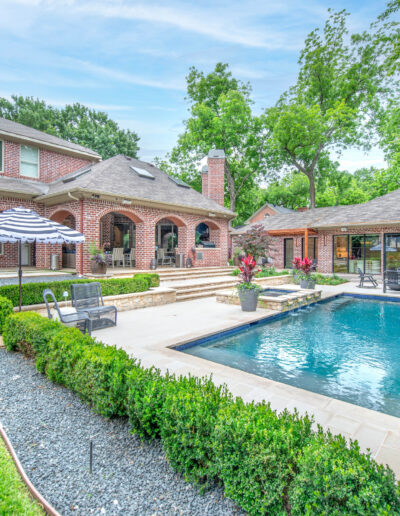 A spacious backyard features a rectangular swimming pool, lounge chairs, a large umbrella, manicured hedges, and trees. A brick house and a pool house are visible in the background under a partly cloudy sky.