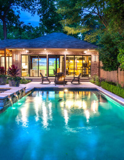 A modern backyard with a glowing swimming pool, patio seating, lush greenery, potted plants, and a house in the background lit warmly at dusk.