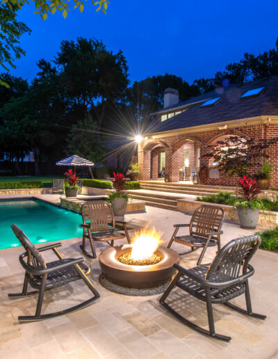 A backyard features a lit fire pit surrounded by chairs on a stone patio, next to a swimming pool. The brick house in the background is illuminated at dusk, with landscaping and trees around the yard.