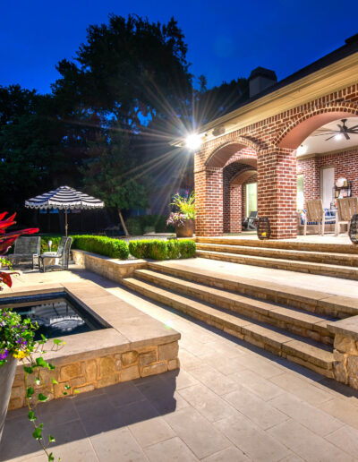 A well-lit backyard patio at dusk features brick arches, steps, a pool with potted plants, and an outdoor dining area with an umbrella surrounded by trees.